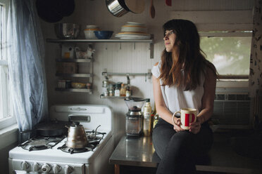 Thoughtful woman holding mug while sitting on kitchen counter - CAVF23921