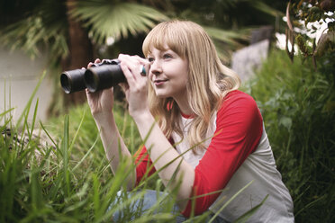 Smiling teenager looking through binoculars while standing by plants at park - CAVF23937