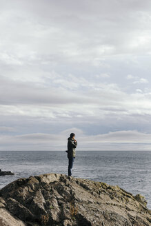 Side view of man standing on rock formation by sea against cloudy sky - CAVF24026