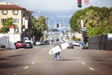 Rear view of man holding surfboard and standing on street - CAVF24152