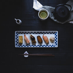 Draufsicht auf verschiedene Sushi, die in einem Teller mit Stäbchen und grünem Tee auf einem schwarzen Tisch angeordnet sind - CAVF24324