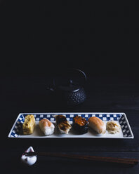 High angle view of various sushi arranged in plate with chopsticks and teapot on table against black wall - CAVF24325