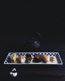 High angle view of various sushi arranged in plate with chopsticks and teapot on table against black wall - CAVF24325