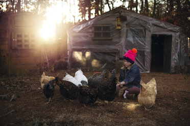 Girl looking at hens while sitting on field against house - CAVF24384