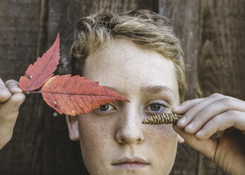 Nahaufnahme eines Jungen, der Herbstblätter und einen Tannenzapfen gegen Holz hält - CAVF24420