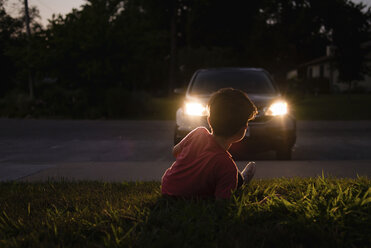Boy relaxing on grassy field while looking at illuminated car at dusk - CAVF24426