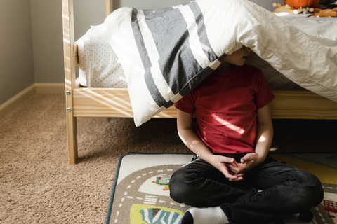 Junge, der mit einer Decke auf dem Kopf an einem Etagenbett sitzt, lizenzfreies Stockfoto