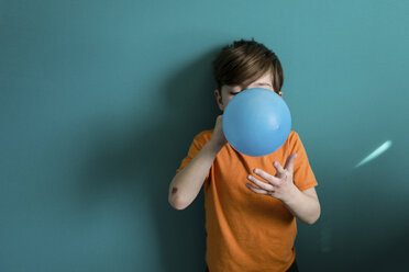 Boy blowing balloon while standing against blue wall at home - CAVF24434