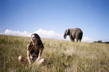 Thoughtful woman sitting on landscape with elephant in background - CAVF24599