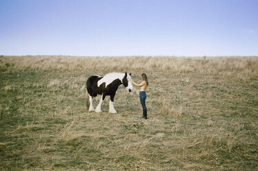 Seitenansicht einer barbusigen Frau, die ein Pferd auf einem grasbewachsenen Feld streichelt, gegen einen klaren Himmel - CAVF24605