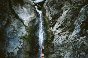 Rear view of naked woman with arms raised standing in front of waterfall - CAVF24608