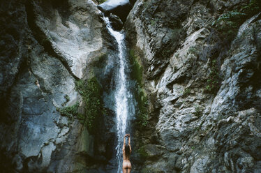 Rückansicht einer nackten Frau mit erhobenen Armen, die vor einem Wasserfall steht - CAVF24608