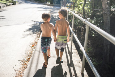 Rear view of shirtless brothers holding hands while walking on footpath by railing during summer - CAVF24689