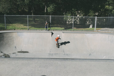 Junge steht, während sein Bruder auf einer Sportrampe im Park Skateboard fährt - CAVF24699