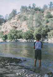 Rear view of boy fishing while standing in lake against mountains - CAVF24708