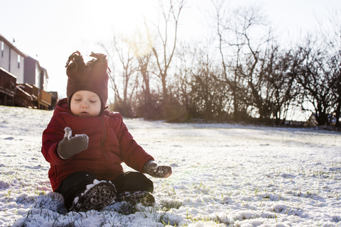 Junge in warmer Kleidung auf einem schneebedeckten Feld im Hinterhof sitzend, lizenzfreies Stockfoto
