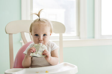 Porträt des niedlichen Baby-Mädchens, das Apfelmus aus der Packung isst, während es auf dem Hochstuhl zu Hause sitzt - CAVF24815