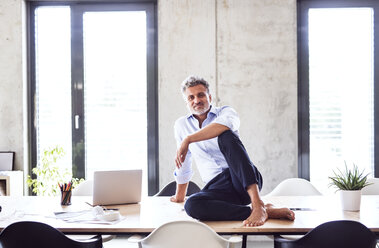 Portrait of smiling mature businessman sitting barefoot on desk in office - HAPF02651