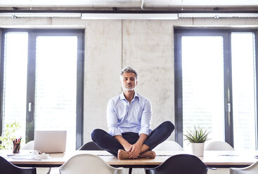 Mature businessman sitting barefoot on desk in office meditating - HAPF02652