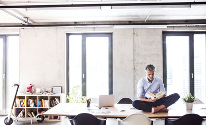 Mature businessman sitting barefoot on desk in office reading a book - HAPF02654