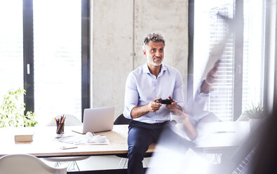 Smiling mature businessman sitting on desk in office using controller - HAPF02657