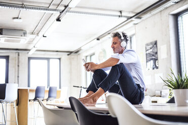 Älterer Geschäftsmann mit Smartphone und Kopfhörern sitzt barfuß auf dem Schreibtisch im Büro - HAPF02659