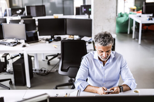 Reifer Geschäftsmann sitzt am Schreibtisch im Büro und benutzt sein Smartphone - HAPF02671