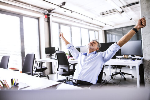 Happy mature businessman sitting at desk in office raising his arms - HAPF02677