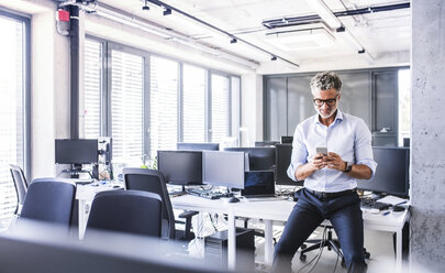 Lächelnder reifer Geschäftsmann sitzt auf dem Schreibtisch im Büro und benutzt sein Smartphone - HAPF02690