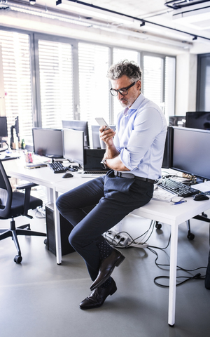 Älterer Geschäftsmann sitzt auf dem Schreibtisch im Büro und benutzt sein Smartphone, lizenzfreies Stockfoto