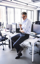 Älterer Geschäftsmann sitzt auf dem Schreibtisch im Büro und benutzt sein Smartphone - HAPF02691