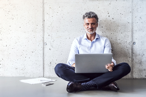 Porträt eines selbstbewussten reifen Geschäftsmannes, der auf dem Boden sitzt und einen Laptop benutzt, lizenzfreies Stockfoto