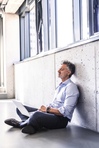 Älterer Geschäftsmann mit Laptop, der auf dem Boden sitzt und sich an die Wand lehnt, lizenzfreies Stockfoto