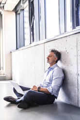 Mature businessman with laptop sitting on the floor leaning against the wall - HAPF02697