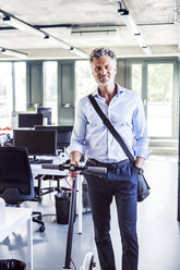 Portrait of smiling mature businessman with scooter in office - HAPF02703