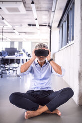 Barefoot mature businessman sitting on floor in office wearing VR glasses - HAPF02714