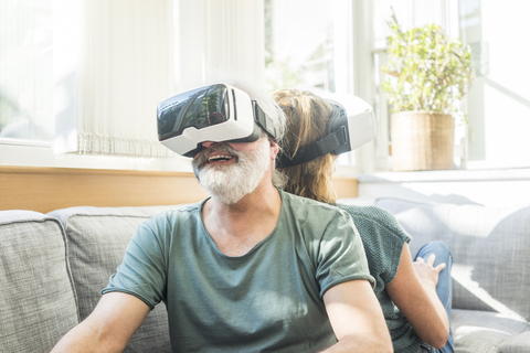 Happy mature couple sitting on couch at home wearing VR glasses stock photo