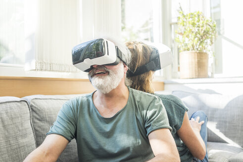 Happy mature couple sitting on couch at home wearing VR glasses - MOEF00937