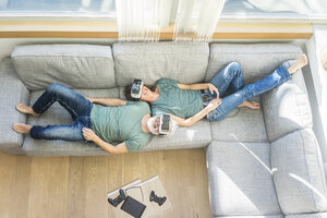 Glückliches reifes Paar, das zu Hause auf der Couch liegt und eine VR-Brille trägt - MOEF00939