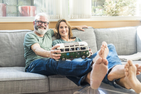 Happy mature couple on couch at home holding minibus model - MOEF00961