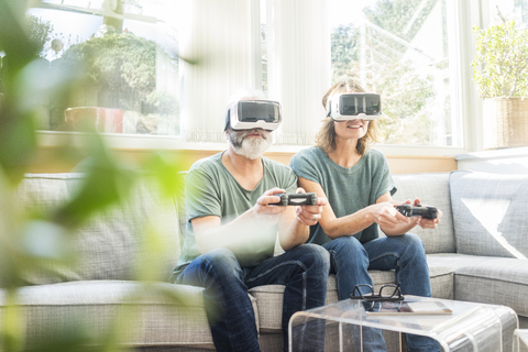 Mature couple sitting on couch at home wearing VR glasses playing video game stock photo