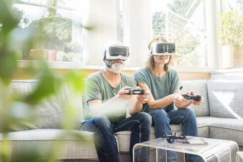 Mature couple sitting on couch at home wearing VR glasses playing video game - MOEF00967