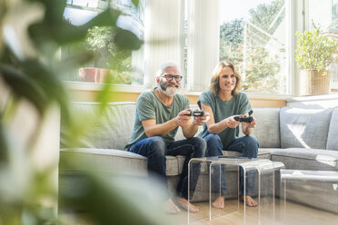 Happy mature couple sitting on couch at home playing video game - MOEF00969