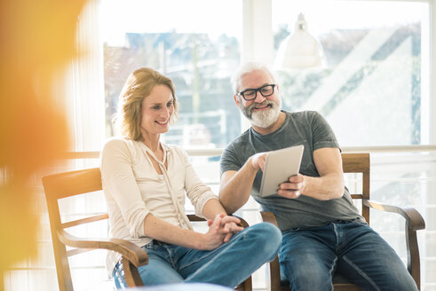 Happy mature couple sitting on chairs at home using tablet - MOEF00992