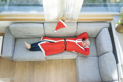 Bearded mature man wearing Santa costume lying on couch in living room stock photo