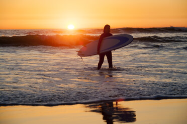 Frau trägt Surfbrett beim Spaziergang am Ufer während des Sonnenuntergangs - CAVF24954