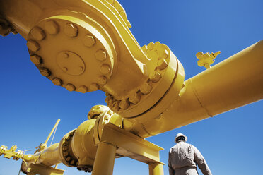 Männlicher Arbeiter steht unter einer gelb gestrichenen Pipeline in einer Ölraffinerie - CAVF25016