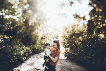 Mother carrying son while standing on dirt road at forest - CAVF25083