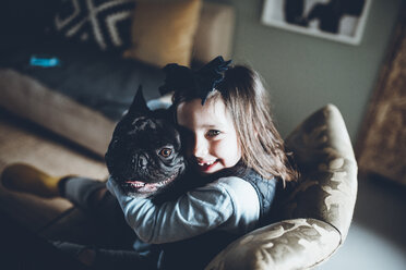 Hochwinkliges Porträt eines glücklichen Mädchens, das eine französische Bulldogge umarmt, während es auf dem Sofa zu Hause sitzt - CAVF25093