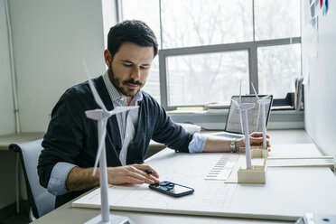 Geschäftsmann benutzt sein Smartphone, während er Windturbinenmodelle auf dem Schreibtisch arrangiert - CAVF25199